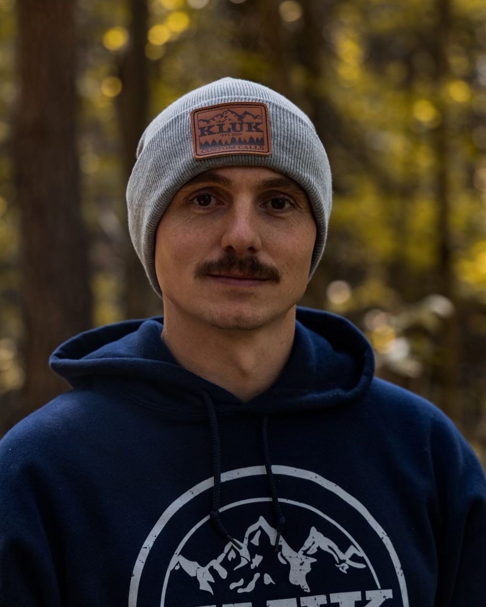 Grey Beanie with leather Kluk logo patch showing off an impeccable stache