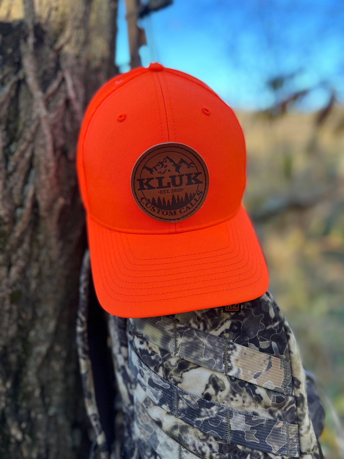 Blaze Orange logo patch hat proped against a tree and a backpack